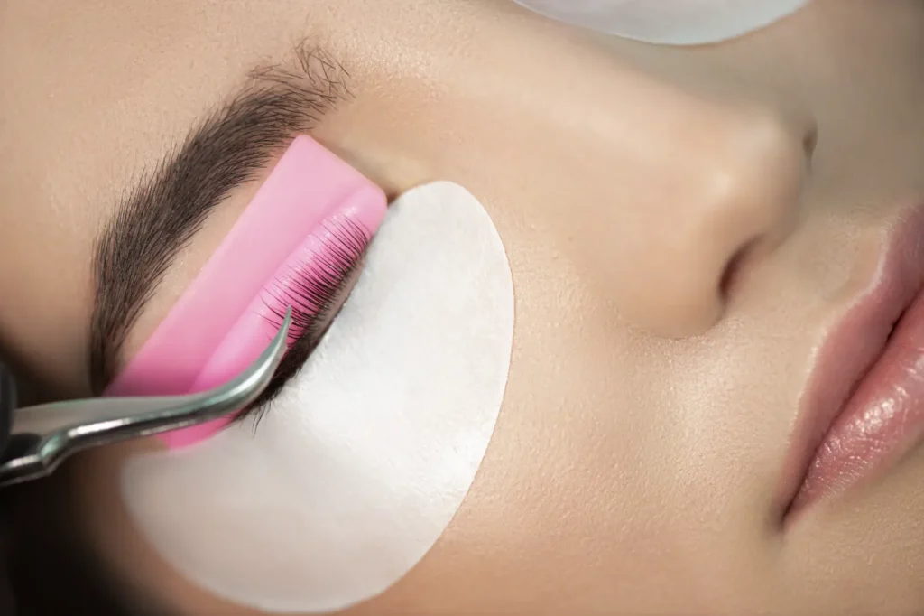 Close-up of tweezers working along a woman's eyelashes with a pink lash tool and under-eye pad.