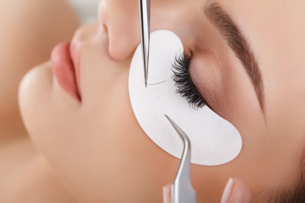 Tweezers hold a single lash near closed eyelashes with a white pad placed beneath the eye.