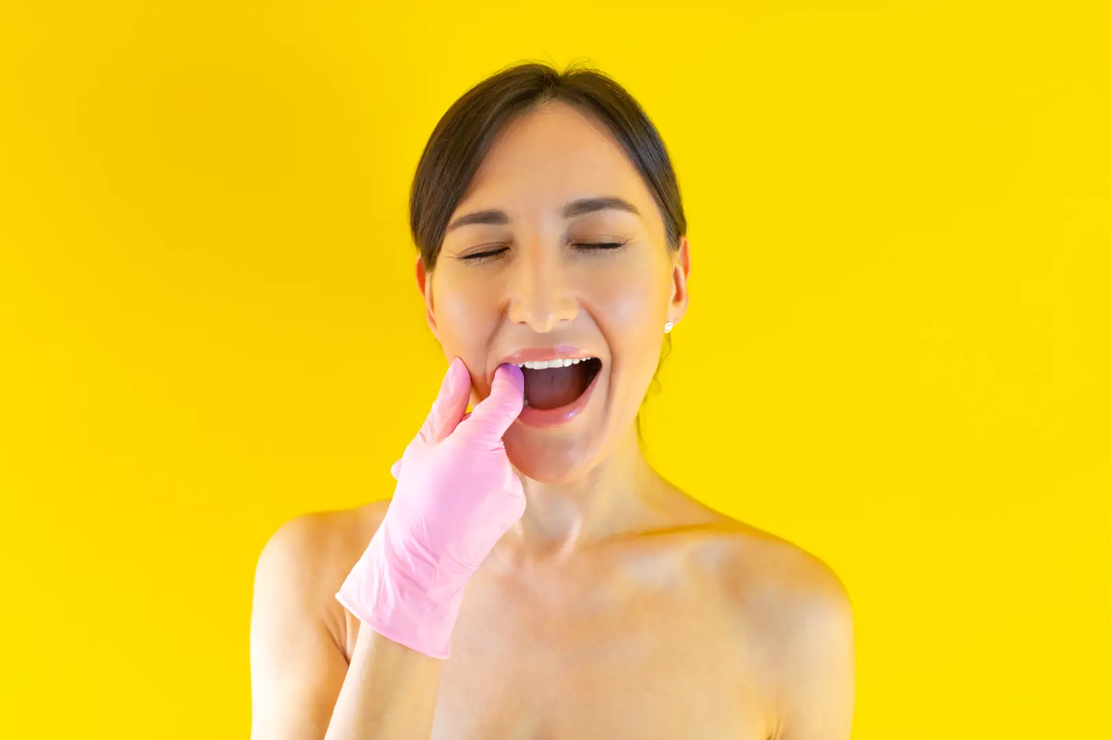 Woman with eyes closed places a pink-gloved finger inside her mouth against a yellow background.